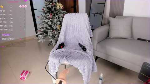 A video showing a woman's feet on a plush grey chair, decorated with pink socks, in a modern living room with a snow-covered Christmas tree and a white sofa.