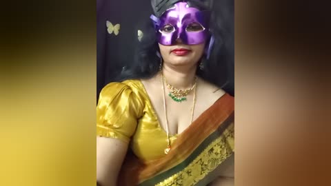 Video of a woman in a purple Venetian mask, gold sari, and green choker, sitting against a black background with a yellow gradient.