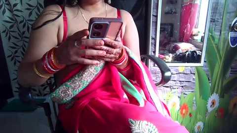 Media: Video of a woman in a bright pink sari, adorned with gold bangles, holding a smartphone in a room with floral wallpaper and colorful decorations.
