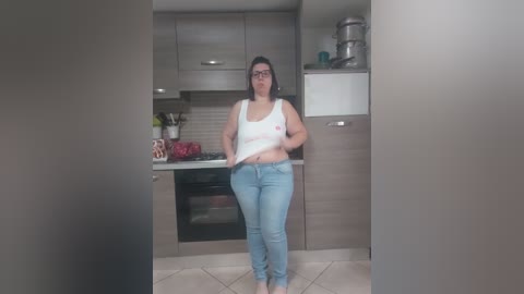 A video of a plus-sized woman with glasses, wearing a white tank top and blue jeans, standing in a modern kitchen with wooden cabinets, stainless steel appliances, and a tiled floor.