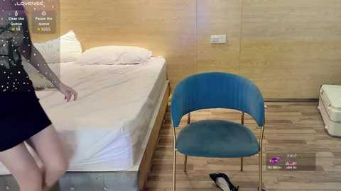 A video of a woman in a black dress, standing near a blue chair in a modern bedroom with wooden walls and a white bed.