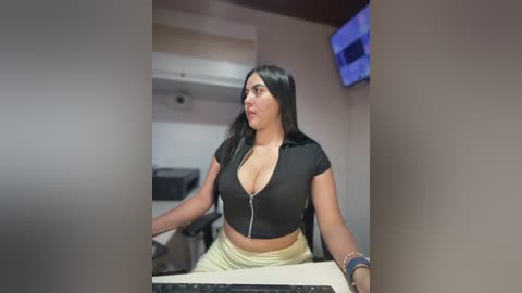 Video of a young woman with long black hair, wearing a low-cut black crop top, sitting at a computer desk in an office.