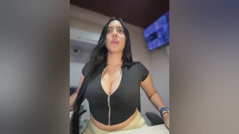 Video of a curvy, light-skinned woman with long black hair, wearing a low-cut black zip-up top and beige pants, making a pouty face in a modern, well-lit room with a TV on the wall.