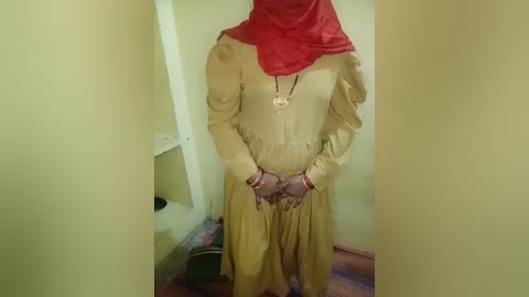 Video of a person wearing a beige long dress with ruffled sleeves, a red headscarf, and gold pendant necklace. They stand in a small, dimly lit room with cream walls and a wooden floor.