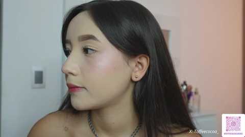 Video of a young Asian woman with straight, long black hair, fair skin, and a slim build. She wears a simple necklace. Background shows a white door and a blurred makeup organizer.