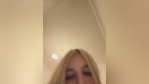 A video of a blonde woman's face reflected in a mirror, with a blurred background. The image has a warm, soft focus, emphasizing her facial features and the mirror's smooth surface.