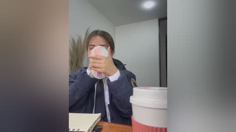 Video of a woman in a dark blue jacket and glasses, holding a colorful mask, sitting at a desk with a white coffee cup and notebook. Background shows a modern office with white walls and a potted plant.