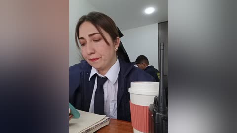 Media: A video of a young woman with fair skin and brown hair, dressed in a dark blue blazer over a white shirt and tie, holding a coffee cup, seated at a table.