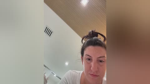 Video of a woman with dark hair in a bun, wearing a white top, looking down, in a modern room with beige walls and wooden ceiling panels.