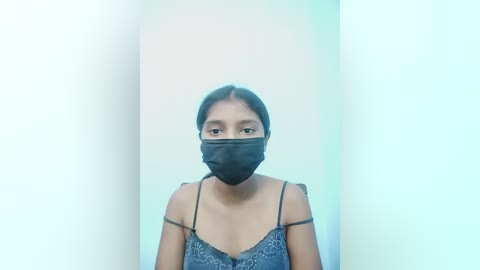Video of a young South Asian woman with medium brown skin, wearing a black mask and blue lace camisole. She stands in front of a bright, gradient background with soft, pastel colors.