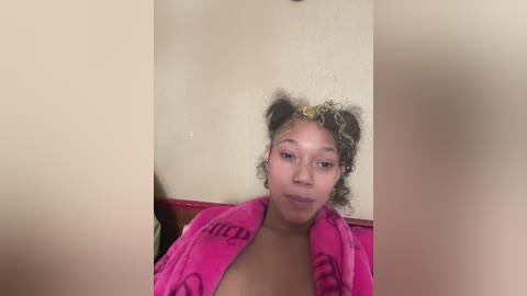 A video of a young Black woman with curly hair in two buns, wearing a pink towel, standing against a beige wall.