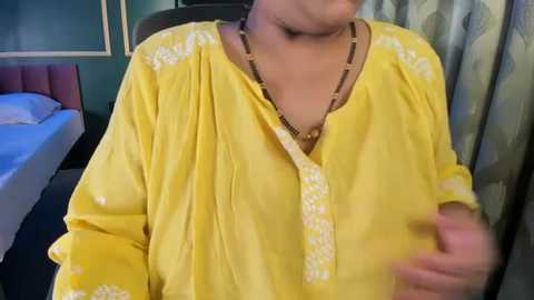 A video of a person wearing a bright yellow kurta with intricate embroidery, a long necklace, and a blurred background featuring a bed, green walls, and patterned curtains.