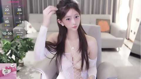Video of a young East Asian woman with long, straight black hair, wearing a white lace off-shoulder top, seated in a modern living room.