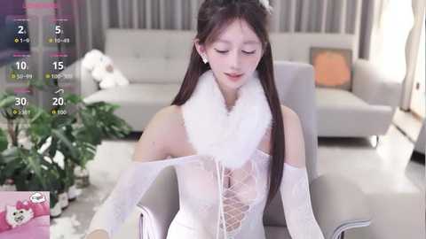 Video of an East Asian woman with long black hair, wearing a white lace off-shoulder dress, fluffy white scarf, and silver earrings, sitting in a modern living room with grey couch, potted plants, and window.