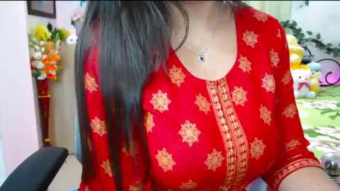 A video of a woman in a red kurta with intricate gold embroidery, long dark hair, and a delicate silver necklace, seated in a room filled with colorful decorations and plush toys.