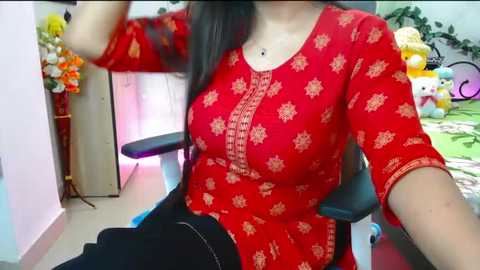 Media: Video of a woman with medium skin tone wearing a red, embroidered kurta with floral patterns, sitting in a black chair in a colorful, playful room filled with toys and plants.