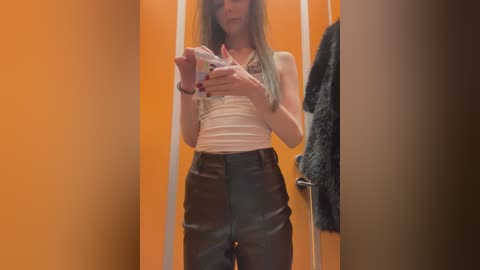 Video of a slender, light-skinned woman with long, straight hair, wearing a white tank top and dark brown leather pants, holding a phone in a brightly lit, orange-walled dressing room, with a dark jacket hanging on a hook.