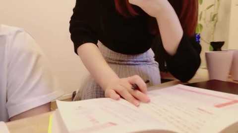 Video of a person in a black shirt and gray skirt, writing on a paper document at a desk, with a white wall and potted plant in the background.