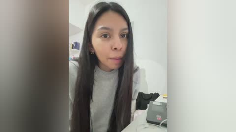 Media: A video of a young woman with long, dark hair and medium-brown skin, wearing a gray t-shirt, sitting in a dimly lit room with cluttered shelves and a laptop.