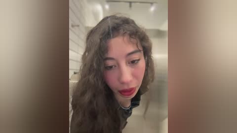 Video of a young woman with curly brown hair, wearing a black jacket, looking down with a slightly surprised expression. The background features a white-tiled wall and a dimly lit room with a ceiling light.