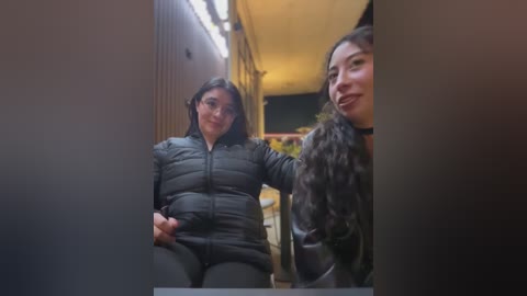 Video of two women with long hair, one wearing a black puffer jacket and the other a leather jacket, standing in a dimly lit, narrow hallway.