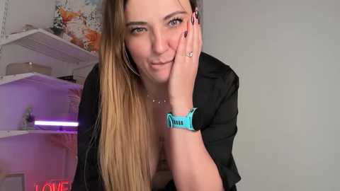 A video of a smiling, fair-skinned woman with long, straight blonde hair, wearing a black top, hoop earrings, and a blue watch. She rests her chin on her hand, in a modern, indoor setting.