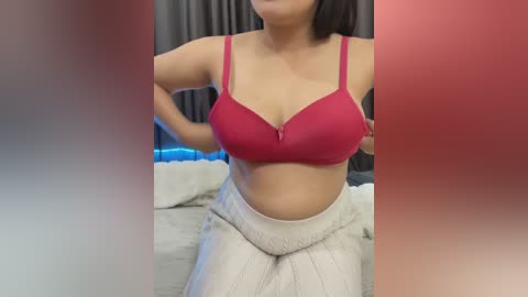 Video of a light-skinned woman with medium-length dark hair, wearing a red bra and white knitted pants, posing in a bedroom with grey curtains in the background.