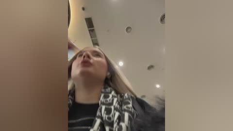 A blurry video of a young woman with light skin and long blonde hair, wearing a black and white patterned jacket, standing in a dimly lit, modern indoor space with recessed ceiling lights.