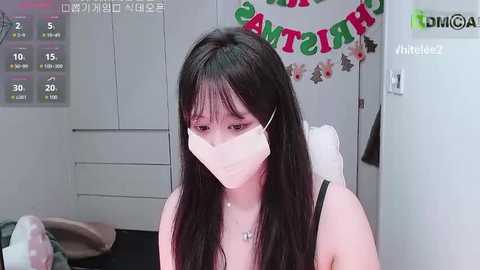 Video of a young Asian woman with long black hair and pale skin, wearing a white surgical mask, black top, and a necklace. She is indoors, looking at a camera, with a calendar and \"Happy Birthday\" banner in the background.