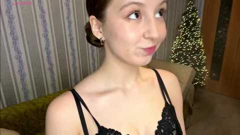 Video of a young woman with light skin, brown hair in a bun, wearing a black lace bra, standing indoors beside a decorated Christmas tree and patterned wallpaper.