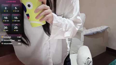 Media: Video of a woman with long black hair, wearing a white blouse, taking a selfie in a room with a white chair and blue cushion.