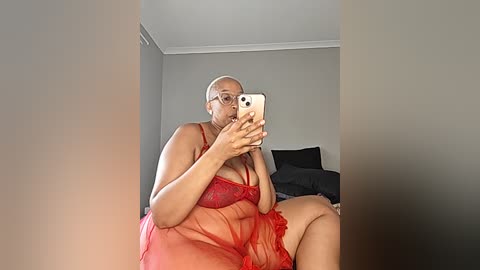 Video of a plus-sized Black woman with short hair and glasses, wearing a sheer red lingerie dress, sitting on a bed with black pillows in a simple, gray-walled room.