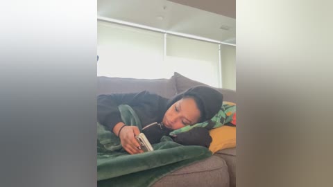 A video of a person lying on a gray couch, wearing a black hoodie and a green blanket, reading a book, with a modern, minimalist room in the background.