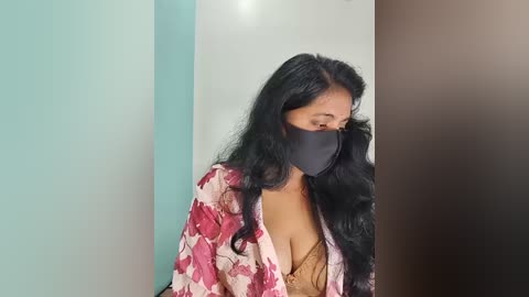 Media: Video of a woman with long black hair, wearing a floral-patterned robe, black mask, and beige lace bra, against a light teal, white, and dark brown vertical wall background.