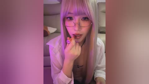 Video of an Asian woman with long, straight platinum blonde hair, wearing glasses, holding a lollipop. She is dressed in a white shirt and black lingerie, posing seductively on a couch.