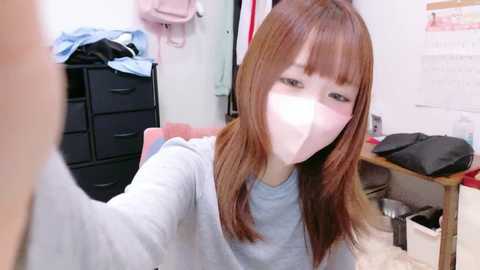 A video of a young Asian woman with straight brown hair and a surgical mask, wearing a light gray shirt, taking a selfie in a cluttered room with black drawers, a pink bag, and clothes.