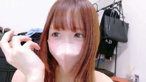 Video of a young Asian woman with long, straight, auburn hair, wearing a face mask, holding a brush, in a cluttered bedroom with black bags and clothes hanging on a rack.