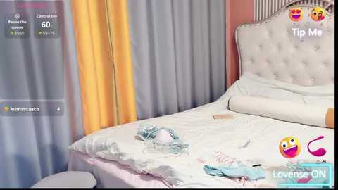 Video of a tidy, modern bedroom with a white, tufted headboard and white bedding. A yellow curtain and pink wall create a bright, cheerful atmosphere. A pair of light blue panties lies on the bed.