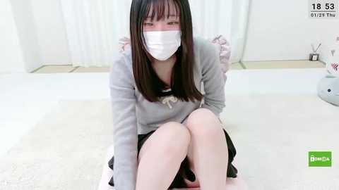 Media: A video of a young Asian woman with long black hair, wearing a face mask and a gray hoodie, sitting on a white floor in a bright, minimalist room with white walls and a small white chair.