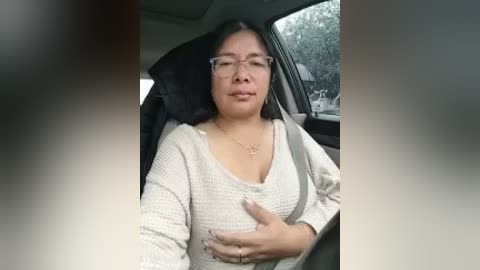 Media: Video of an Asian woman with glasses, medium build, wearing a beige knit sweater, seated in a car with a black seatbelt, looking slightly uncomfortable.