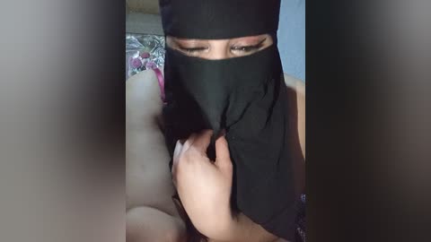 A video of a person wearing a black niqab, partially covering their face, with a hand holding the niqab. The background shows a bed with a floral patterned sheet. The scene is intimate and personal.