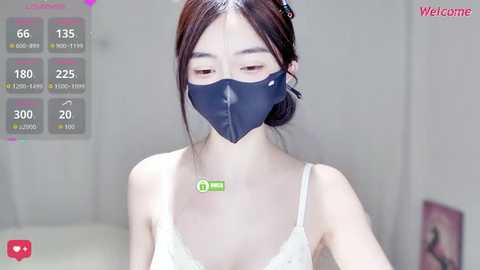 A video of an East Asian woman with fair skin, wearing a white lace top, black face mask, and dark hair in a bun. She stands indoors with a blurred background.