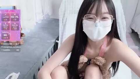 Video of a young East Asian woman with long black hair, wearing a white face mask, glasses, and a pink and white teddy bear onesie, sitting in a white bed.