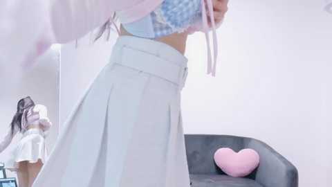 Video of a slender, light-skinned Asian woman in a pastel pink hoodie and white pleated skirt, revealing a blue bra. Background includes another woman in a white shirt and a grey couch with a pink heart pillow.
