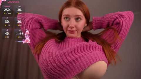 Video of a young woman with fair skin and long, reddish-brown hair in pigtails, wearing a pink knitted sweater that exposes her chest. She has a slight smile and is posed with arms behind her head.