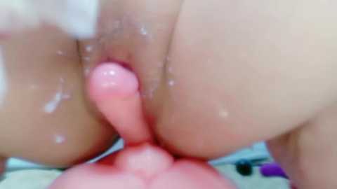 A close-up video of a naked person with light skin, showing a hand holding a pink, smooth dildo inserted into their anus. The background is blurred, likely a bed with soft, colorful bedding.