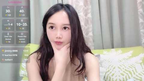 Media: Video of a young East Asian woman with long black hair, fair skin, and a slender build, wearing a green floral-patterned shirt. She's indoors with curtains in the background.