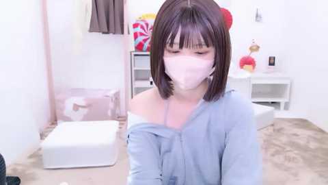 Video of a young Asian woman with short dark hair, wearing a light blue off-shoulder shirt, a white face mask, and a red hair clip. She sits on a white bench in a minimalist, clean bathroom with white walls and a beige floor.