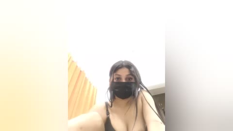 A video of a young woman with long black hair, wearing a black face mask and a black tank top, standing in a brightly lit, minimalist room with yellow and white walls.