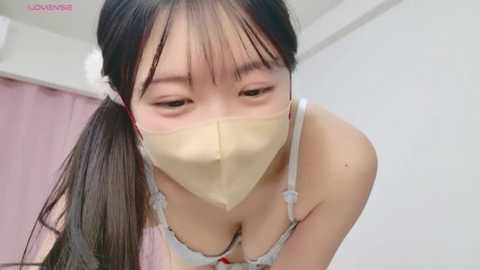 A close-up video of an Asian woman with long black hair in a ponytail, wearing a beige face mask and a revealing white lace bra, leaning forward in a bedroom with white walls and pink curtains.